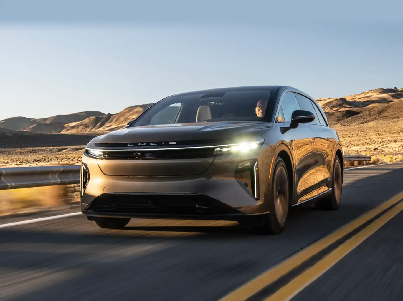 سيارة Lucid Gravity تسير على طريق صحراوي سريع مع إضاءة المصابيح الأمامية، وتتميز خلفية الصورة بالتلال الذهبية تحت السماء الزرقاء.