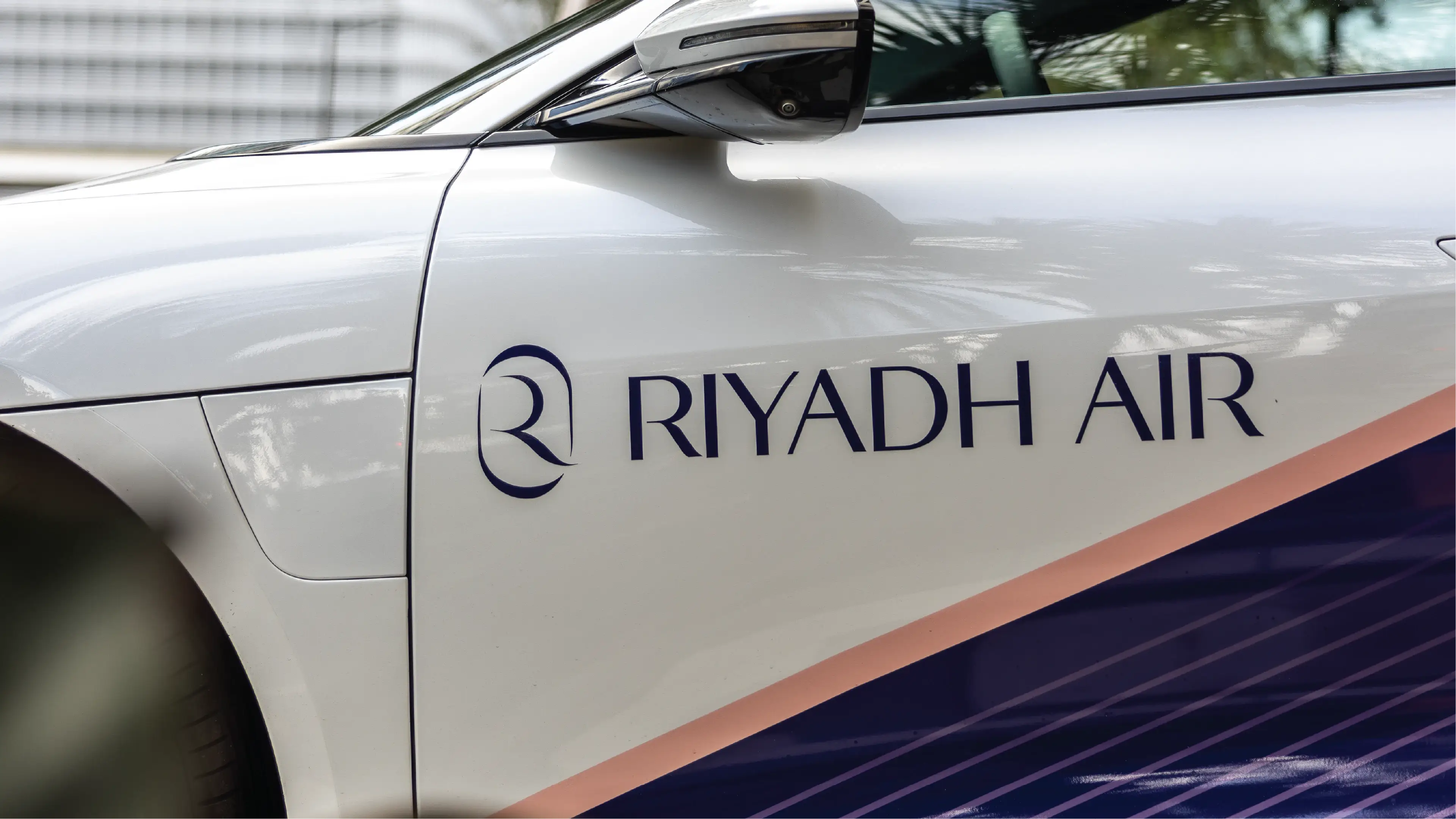 صورة مقربة لمركبة بيضاء تحمل شعار "طيران الرياض" باللون الأزرق الداكن بتصميم أنيق. يبدو اللون أنيقًا واحترافيًا.