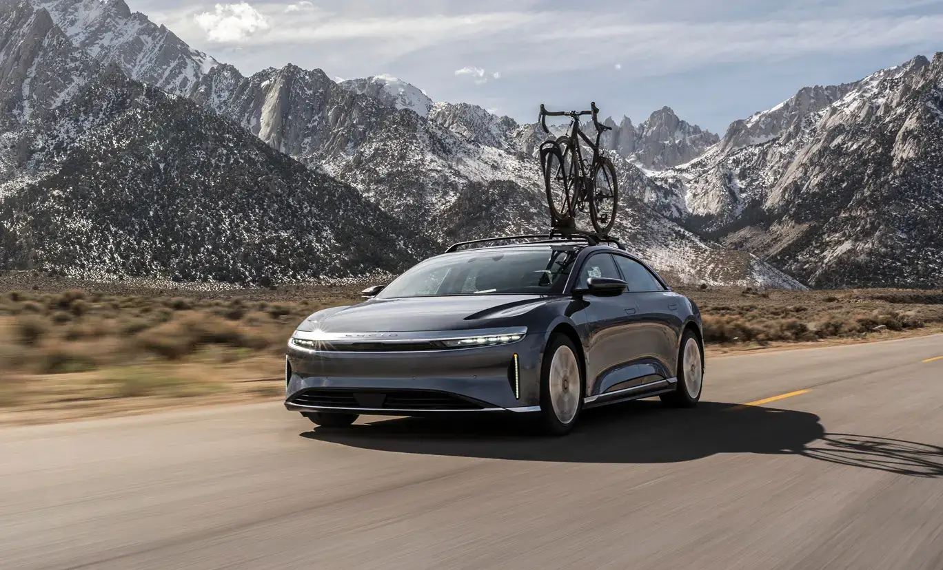 A Lucid Air with a bicycle on the roof rack drives on a road with snow-capped mountains in the background.