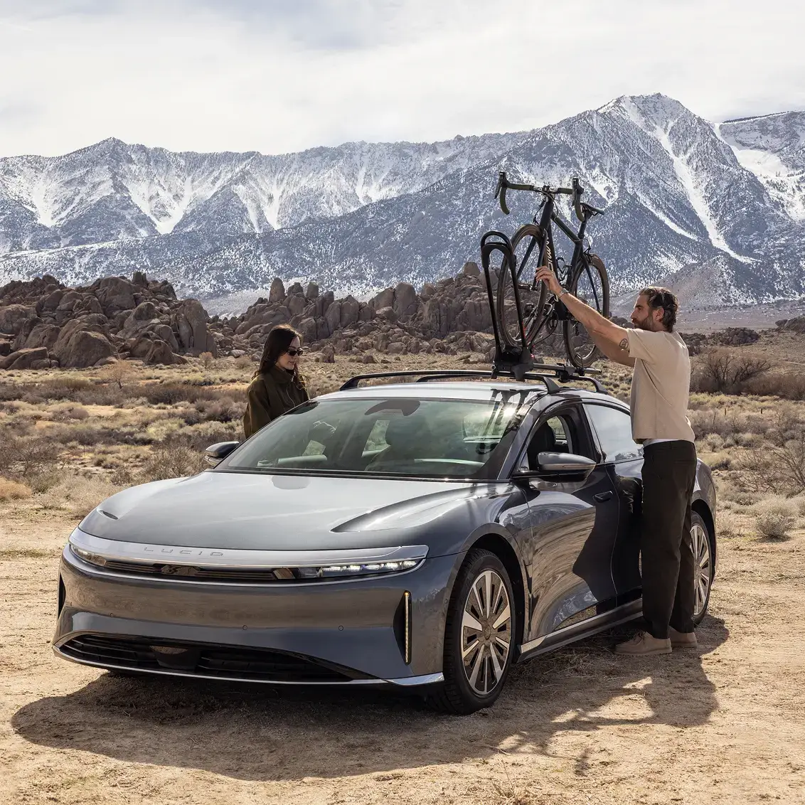 A man loads a bicycle onto a Lucid Air's roof rack in a desert landscape with snow-capped mountains behind.
