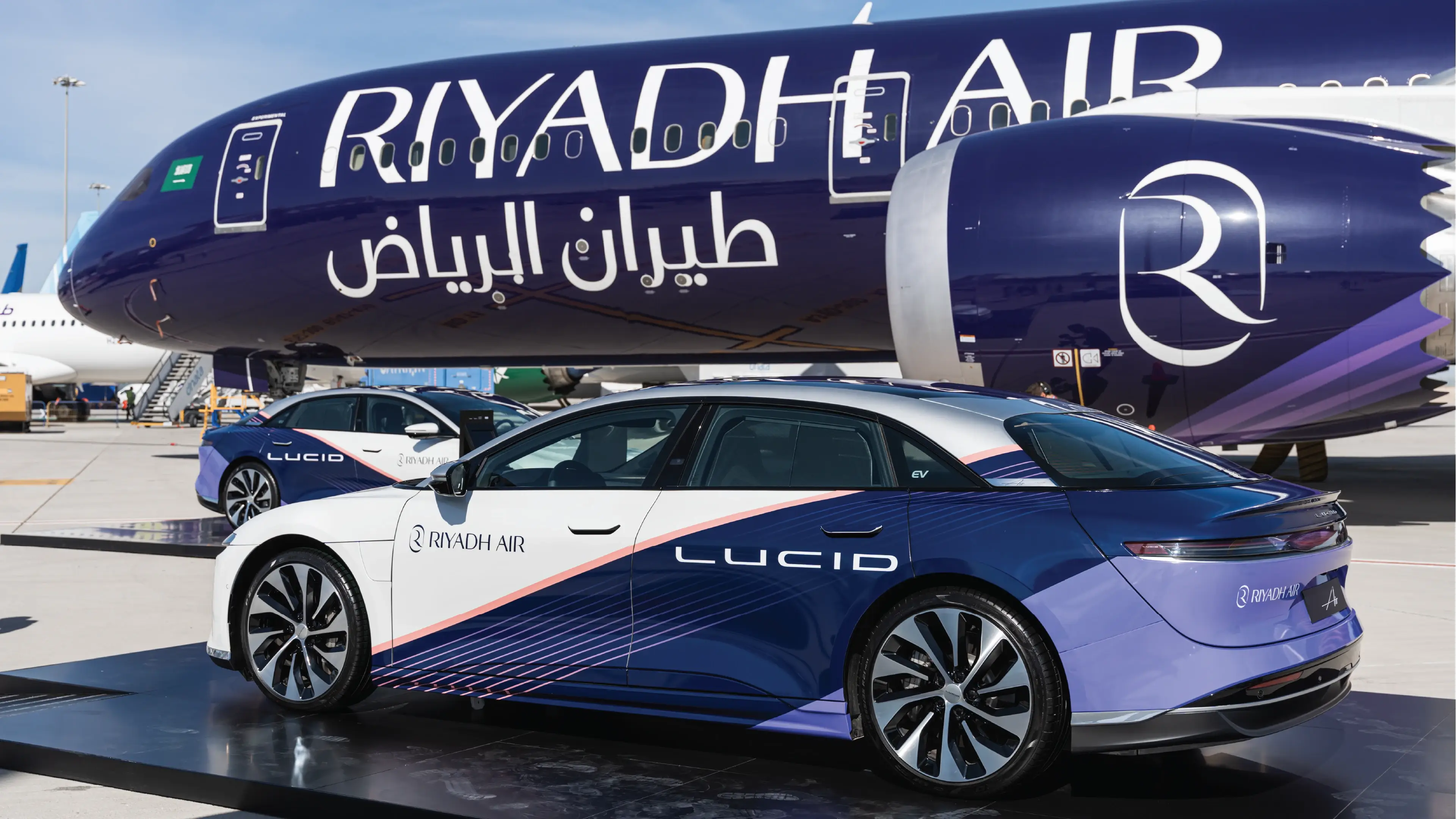 طائرة بنفسجية تحمل علامة طيران الرياض، مع مركبات لوسد الكهربائية بنفس الألوان متوقفة على مدرج المطار تحت سماء زرقاء.