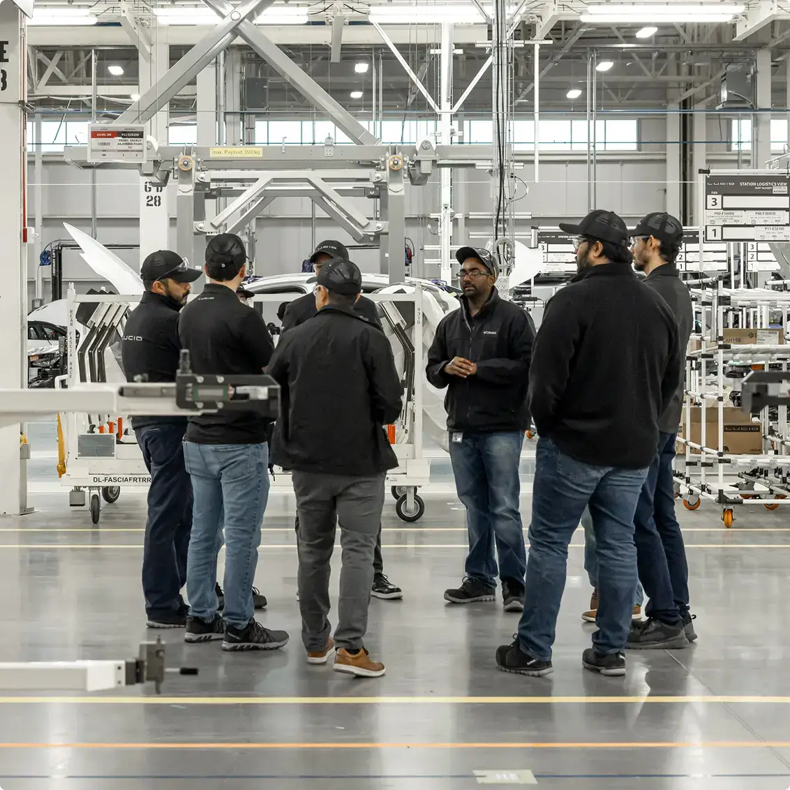 Workers in black jackets gathered in a modern automotive manufacturing facility discussing production components.