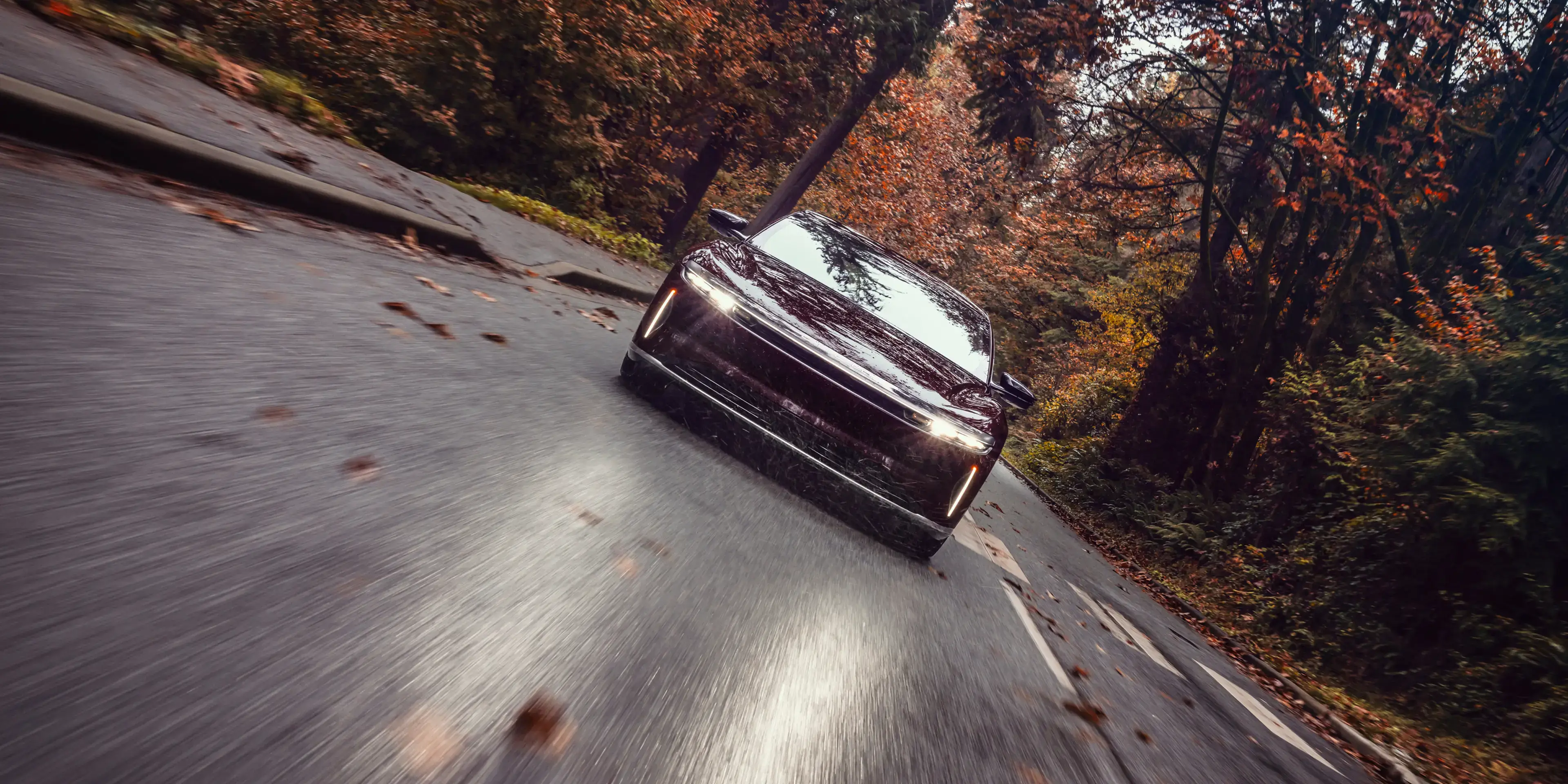 Zenith Red Lucid Air with headlights on driving along a winding autumn road surrounded by orange and red fall foliage.