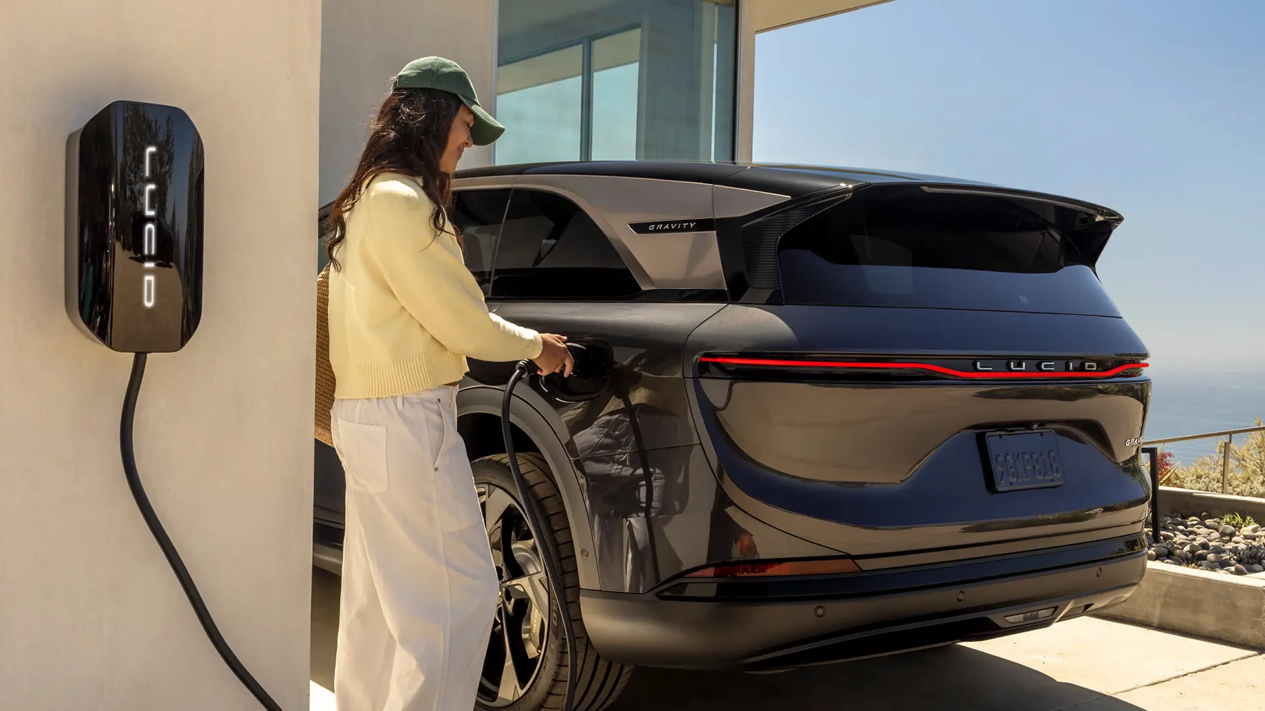 Woman in white outfit plugging a charger into a black Lucid Gravity SUV at a home EV charging station.