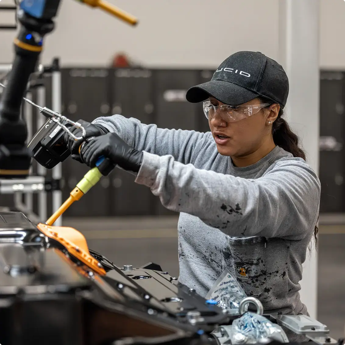 Factory worker in safety glasses and LUCID cap using a power tool on an automotive assembly line.