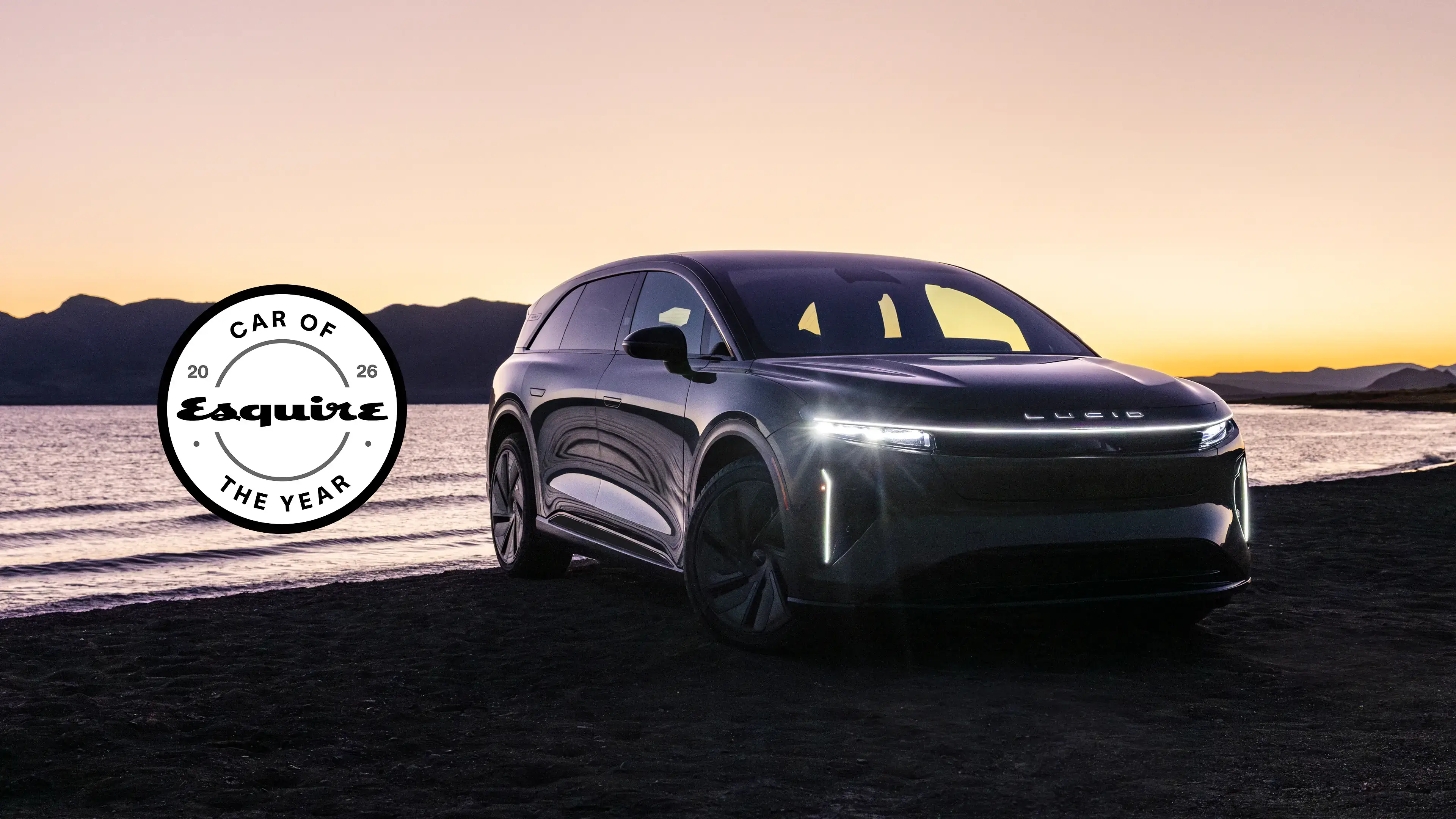 Lucid Gravity parked by water at sunset with mountains in background, headlights illuminated.