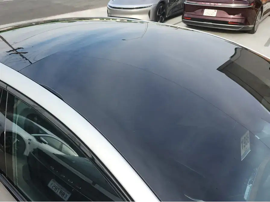 Close-up of a car's black glass roof panel with silver trim, parked alongside other luxury vehicles on a street.