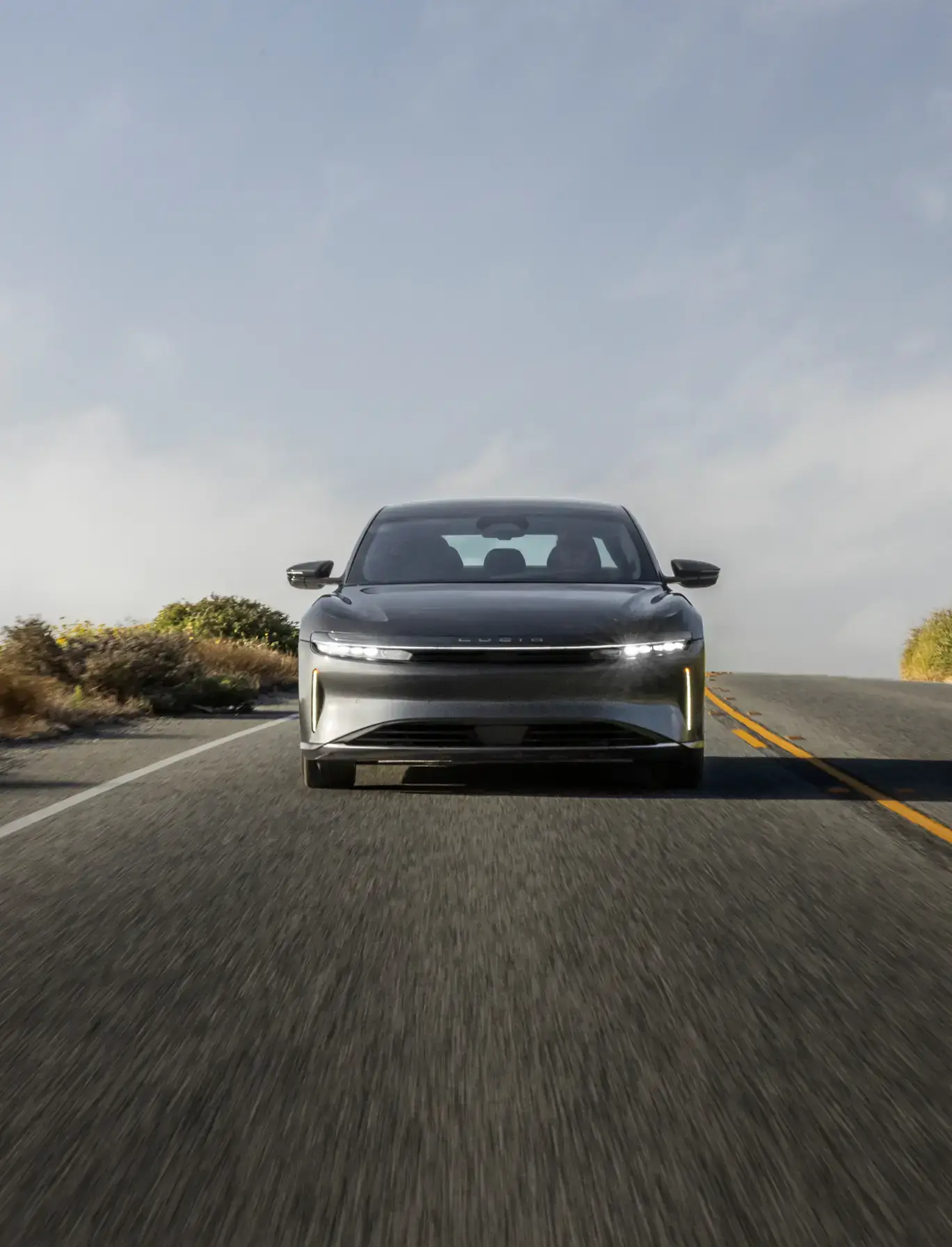 A sleek gray Lucid Air electric sedan driving head-on down a two-lane road with motion blur under a cloudy sky.