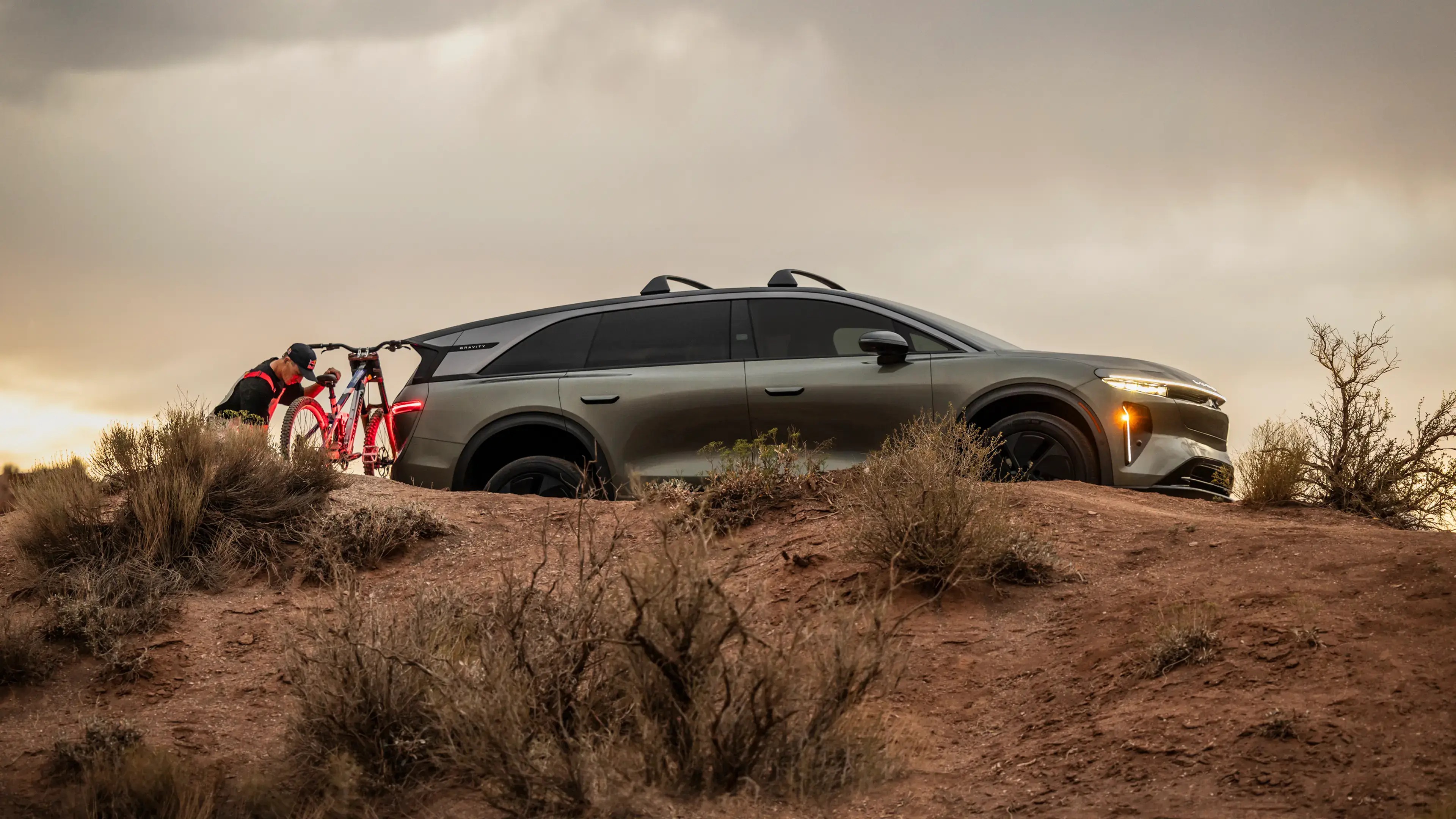 A tan SUV parked on a rocky desert terrain at dusk, with a person loading a red mountain bike onto the rear hitch.