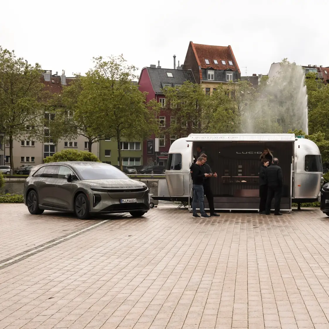 Vue latérale d’un SUV électrique Lucid stationné à côté d’une remorque‑showroom argentée estampillée Lucid, sur une place urbaine colorée, avec des visiteurs à proximité et une fontaine en arrière‑plan.