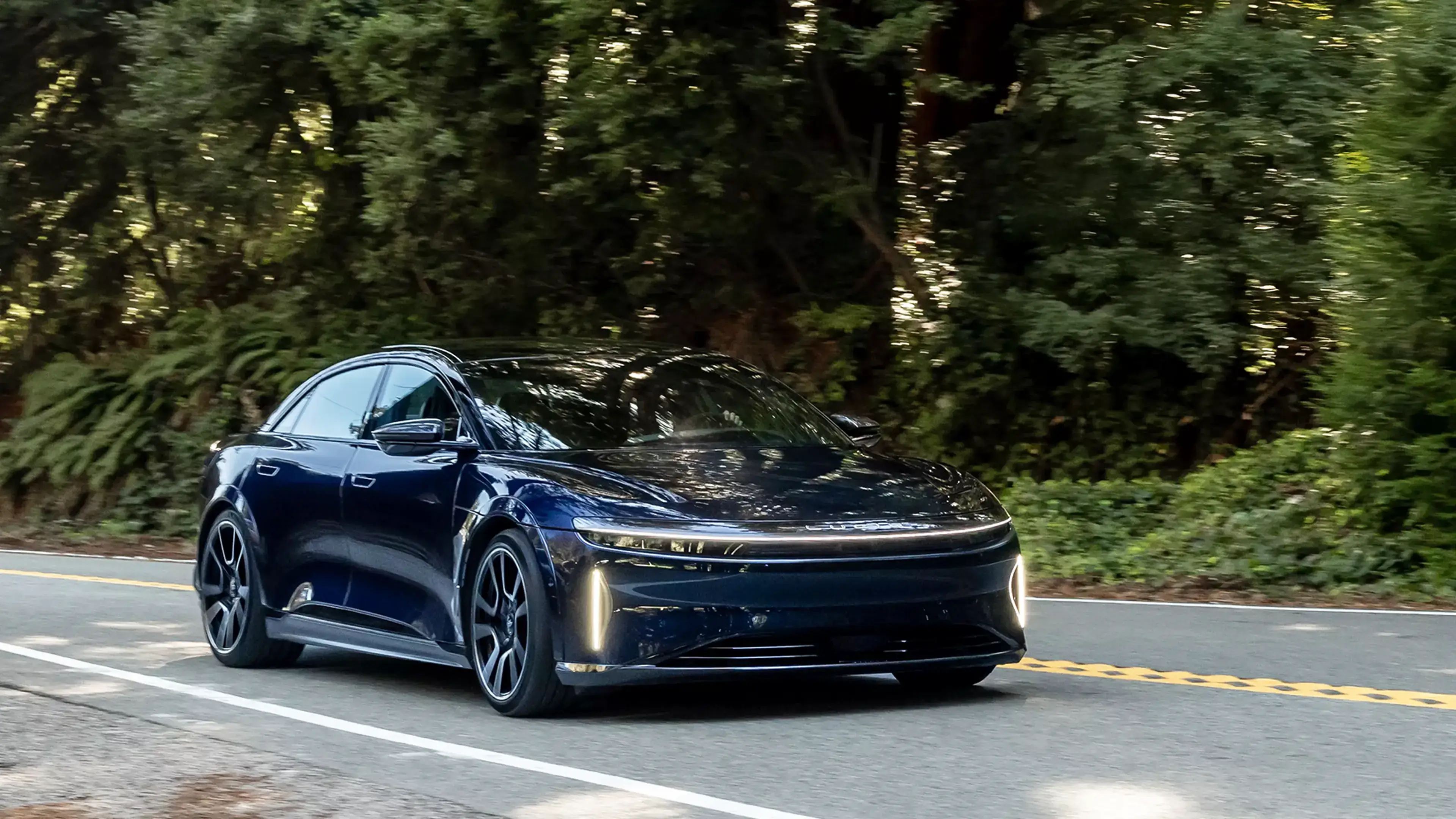 A Lucid Air Sapphire electric sedan driving on a tree-lined road, with illuminated front light strips.