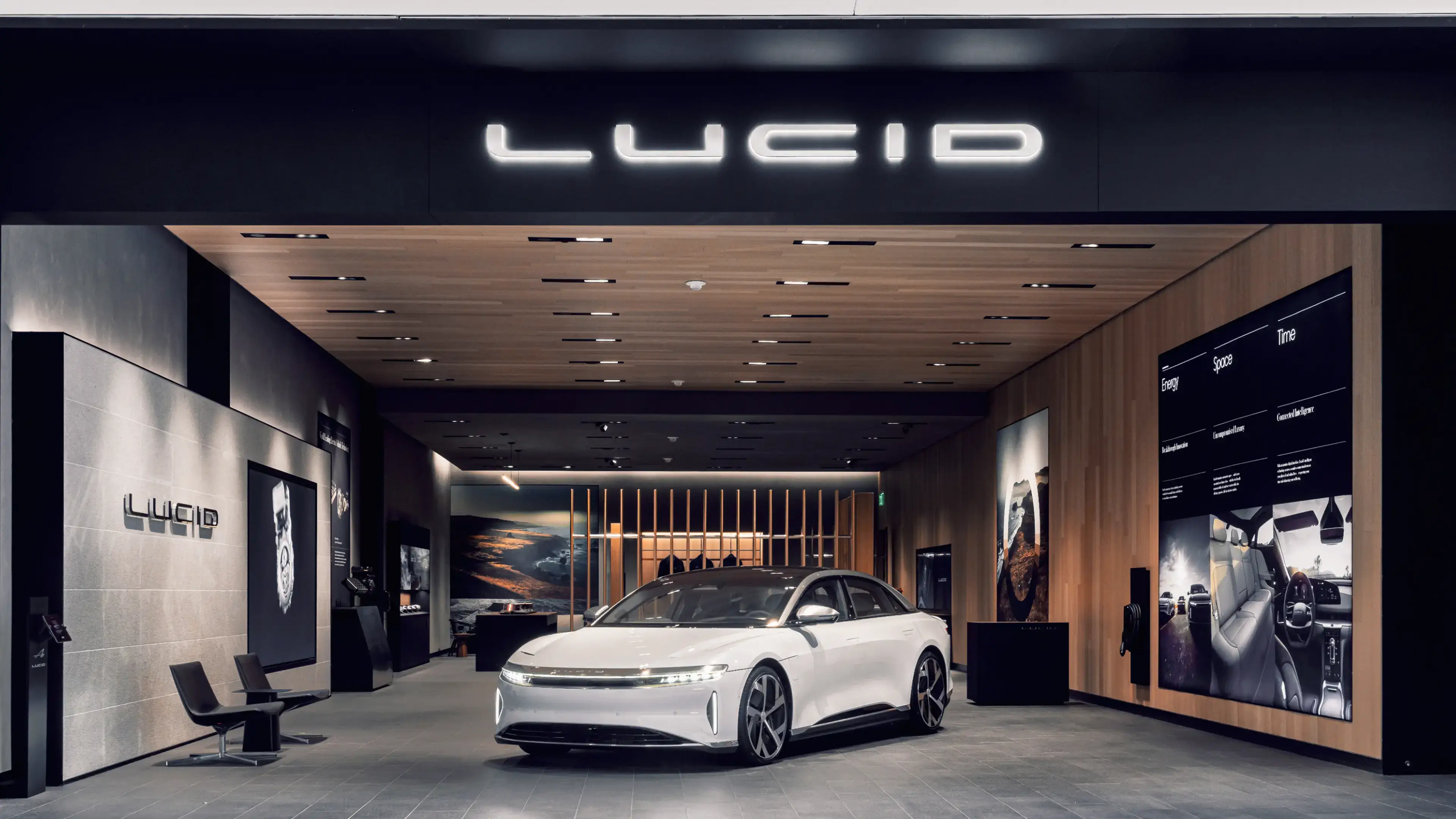 White Lucid electric sedan displayed in modern showroom with wood ceiling, illuminated logo, and digital displays.