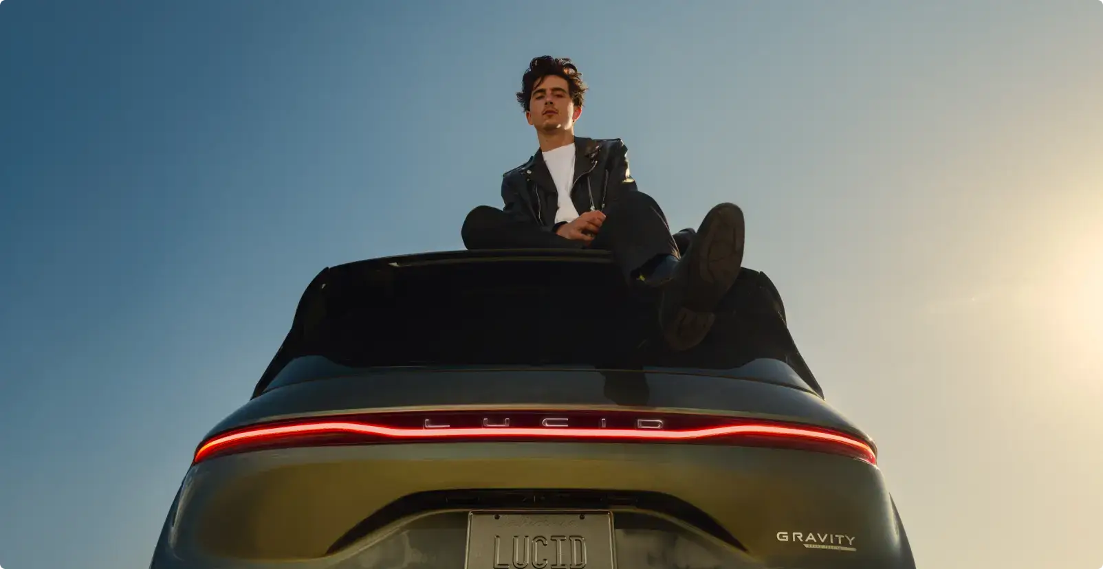 Timothée Chalamet zittend op het dak van de Lucid Gravity tegen een heldere blauwe hemel bij zonsondergang.