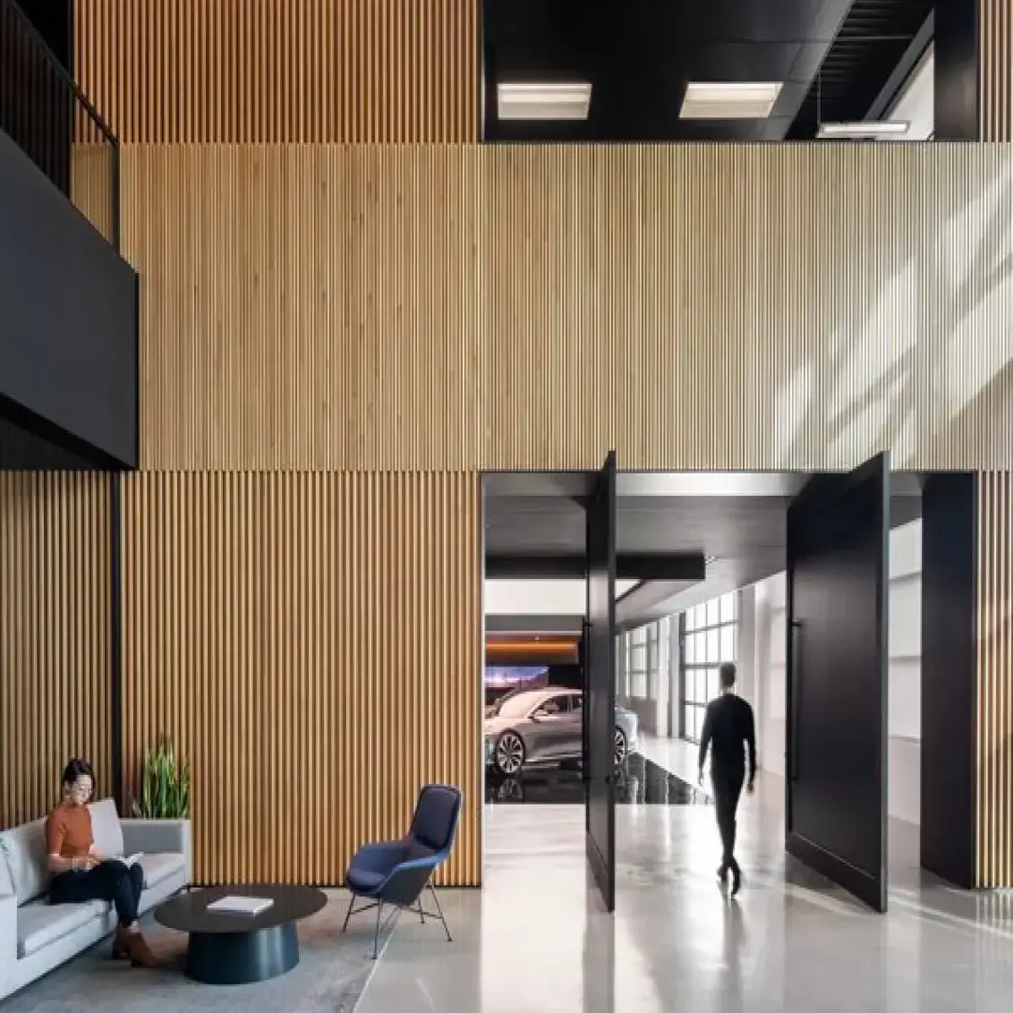 2 people, one sitting in a couch and ine walking away from the camera, are shown in a large space in a Lucid®极速赛车168体彩数据公开查询平台 location. The room mixes wood and black colors, with concrete floors and lots of natural light.