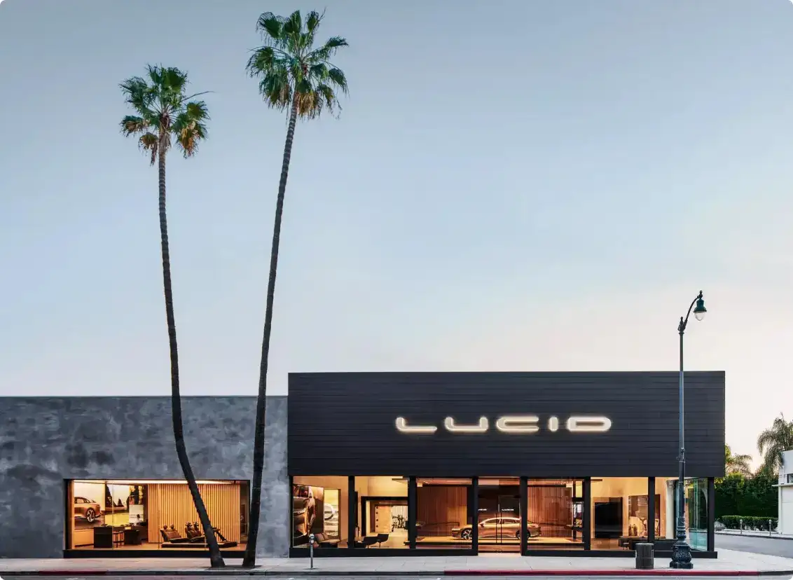 Lucid Motors studio with illuminated signage, modern dark exterior, and two tall palm trees against a twilight sky.