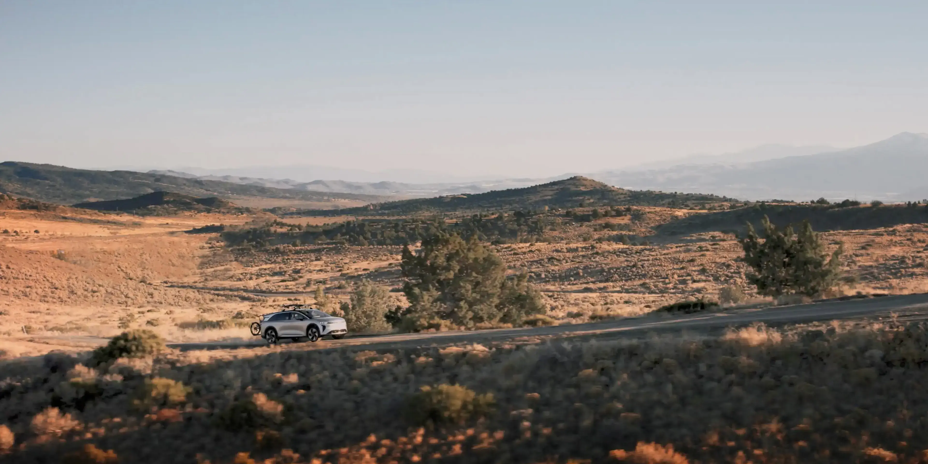The Lucid Gravity diving on on a trail outdoors in a hilly area. The Lucid Gravity has a mountain bike attached to it.