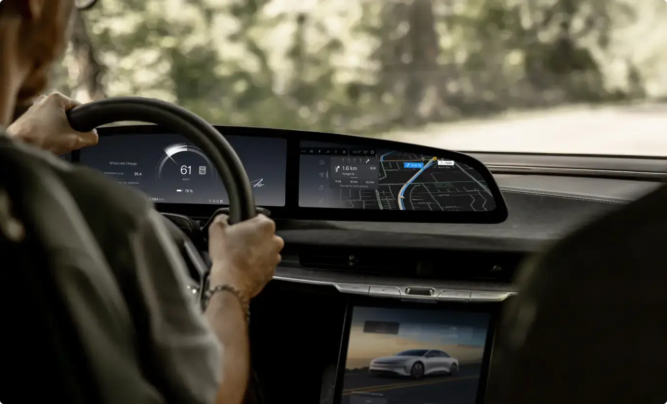 Driver gripping a steering wheel with a wide digital dashboard displaying speed, battery level, and navigation map.
