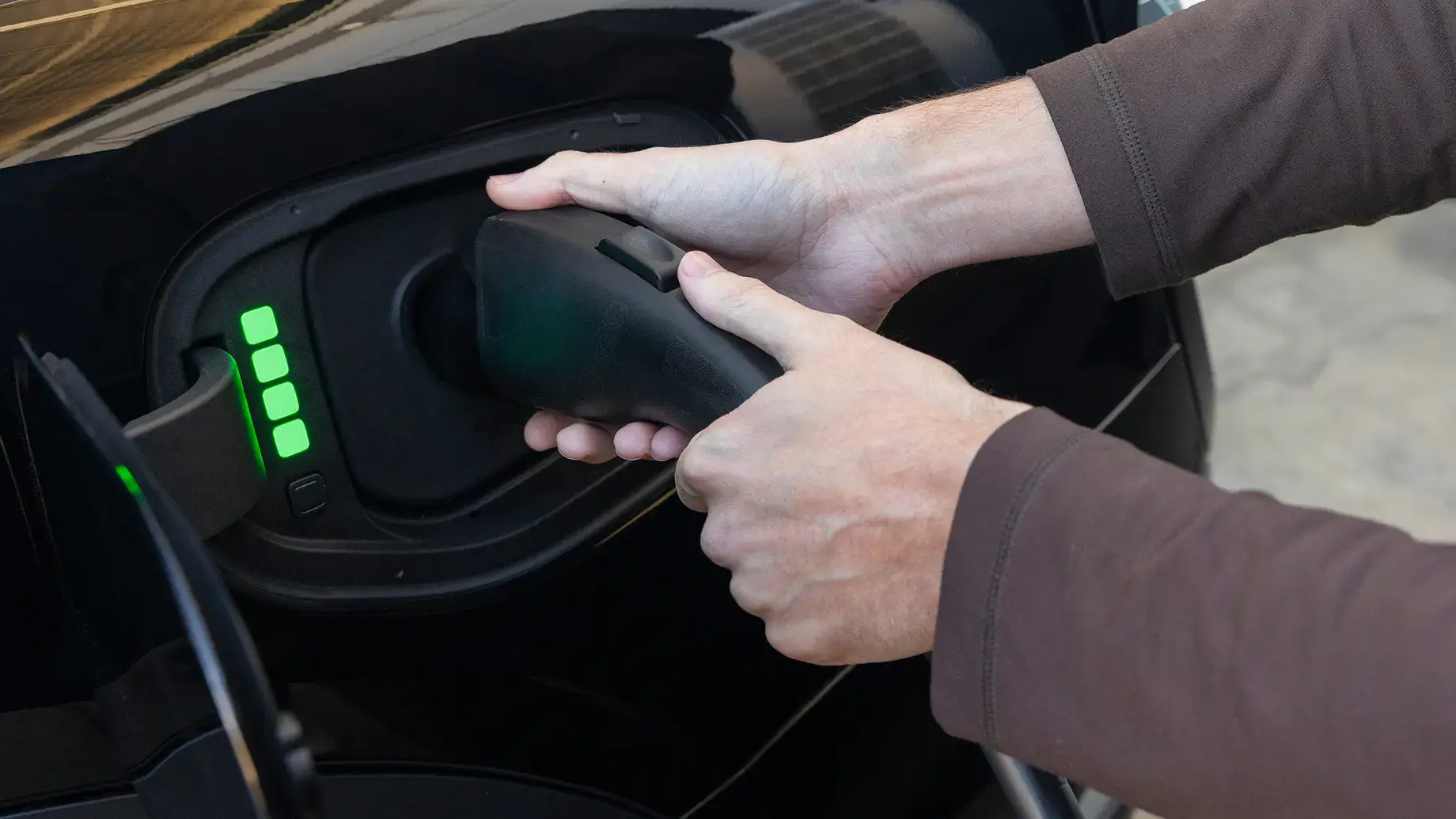 Hands plugging in an electric vehicle charger with green battery indicator lights showing charging progress.