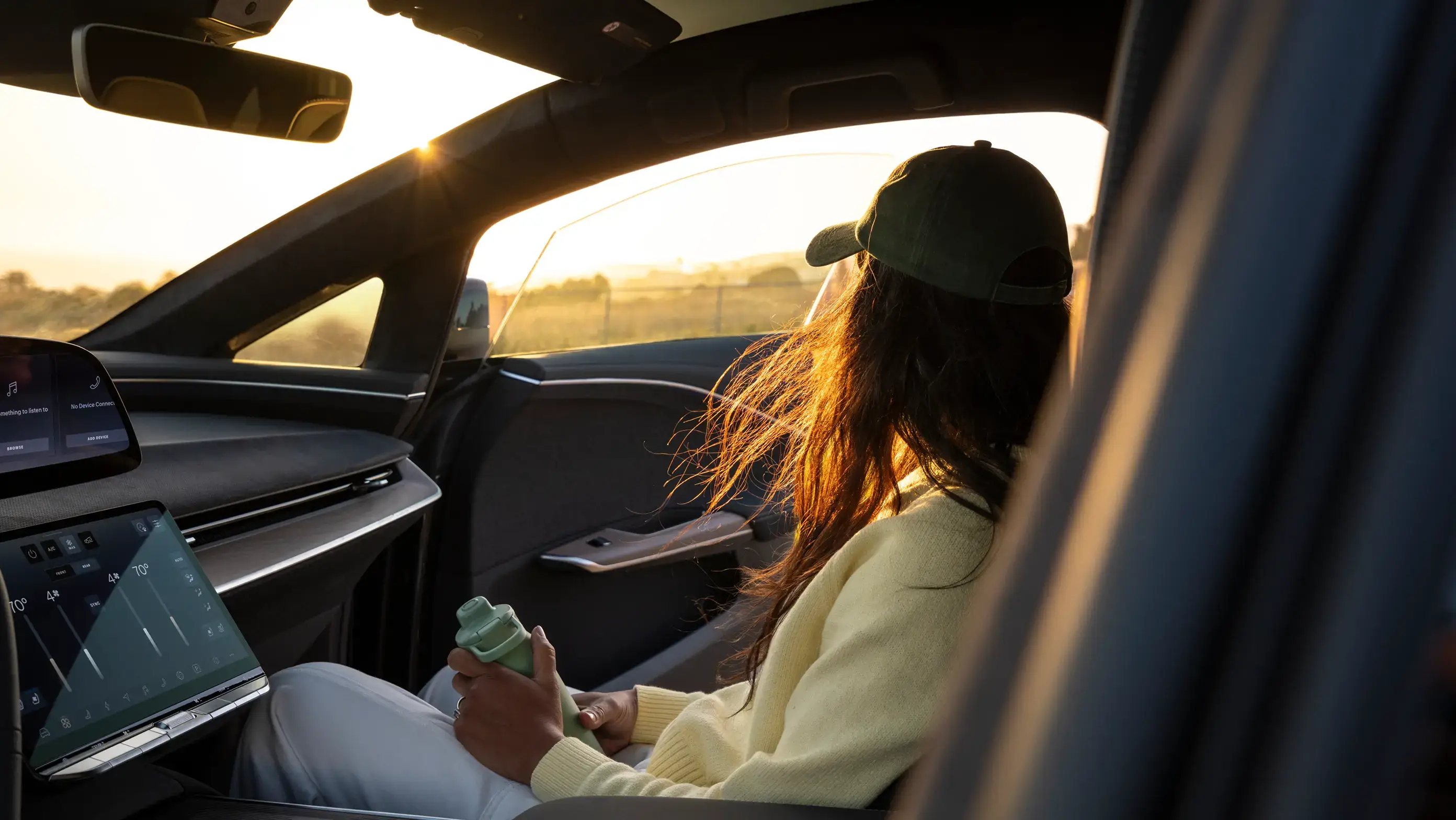 Woman in a yellow sweater and black cap sits in a modern car at sunset, holding a green bottle near a large touchscreen.