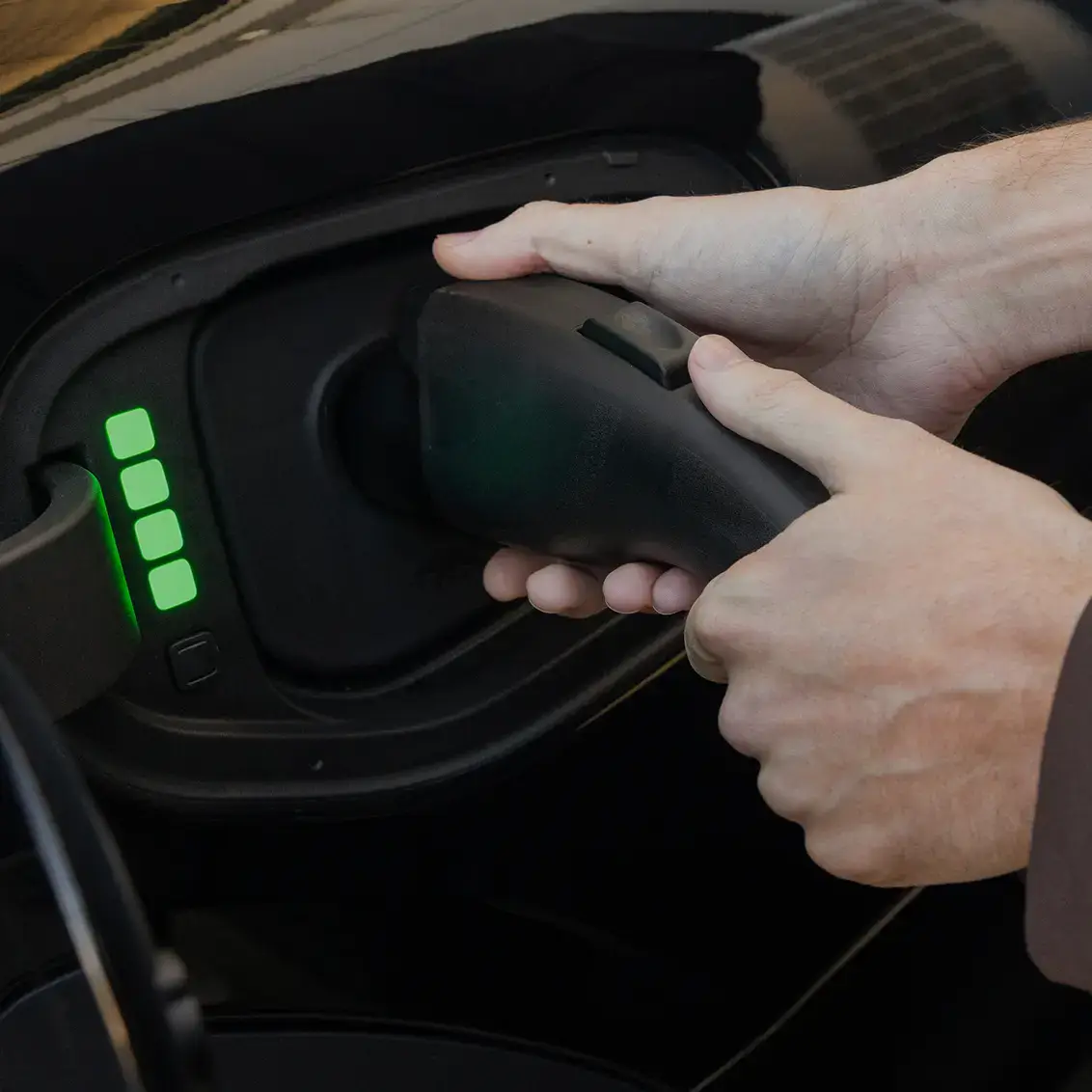 Hands plugging in an electric vehicle charging cable with four green indicator lights showing battery level.