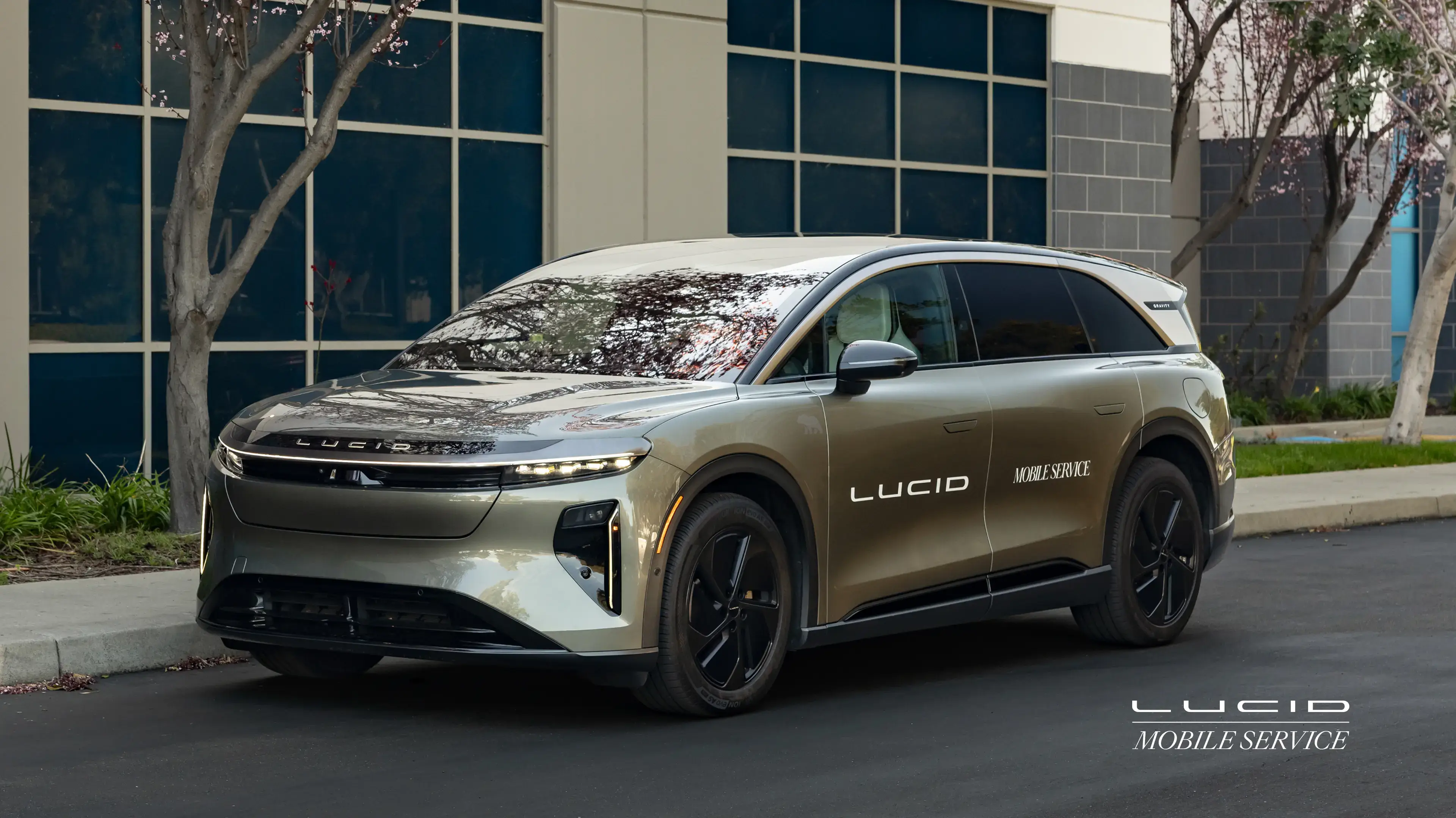 Sleek Lucid Mobile Service electric SUV parked outside modern building with glass facade and flowering trees.