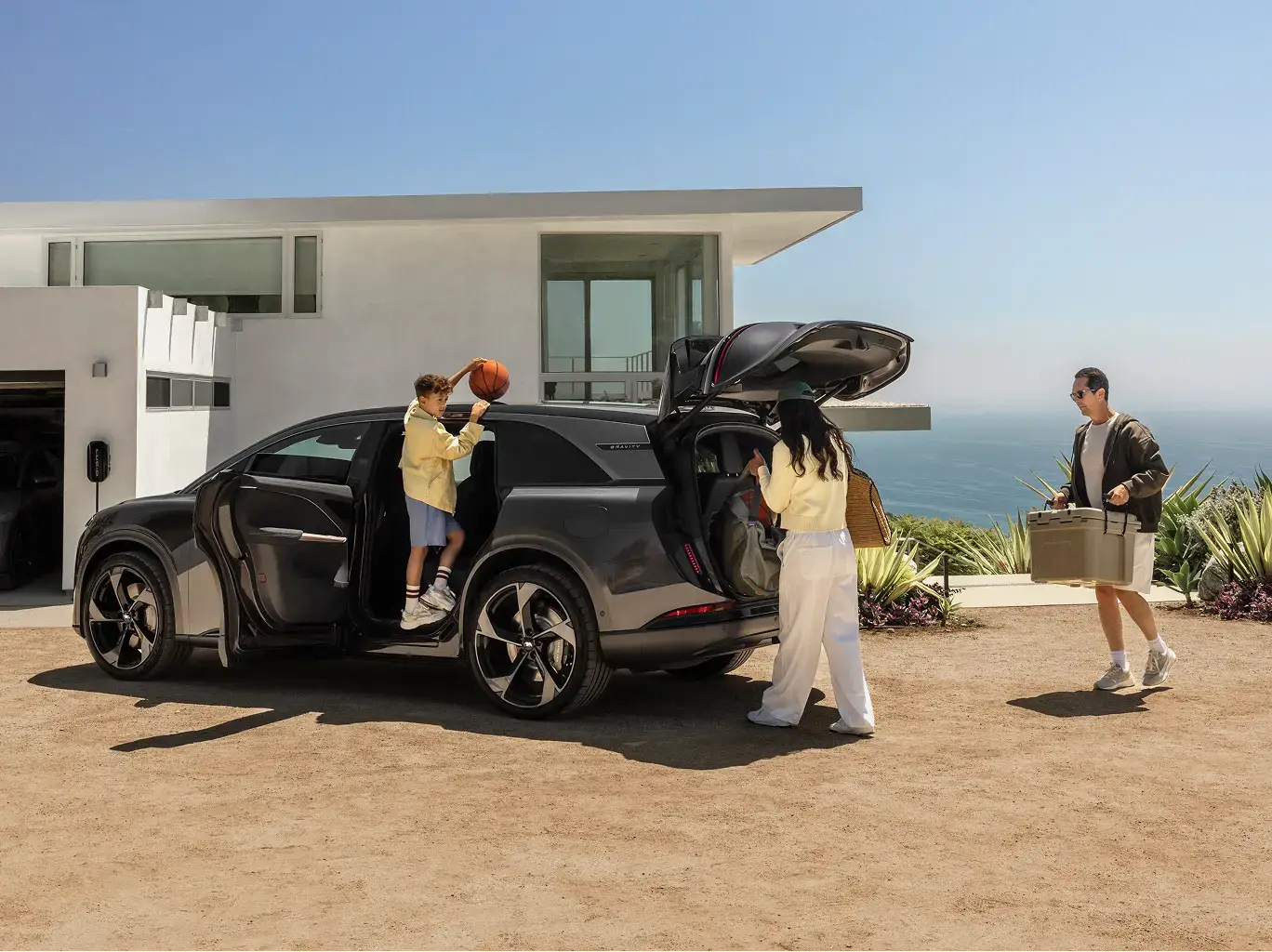 Family loading a Lucid Gravity SUV outside a modern coastal home; a boy holds a basketball, a woman packs the trunk, a man carries a cooler.