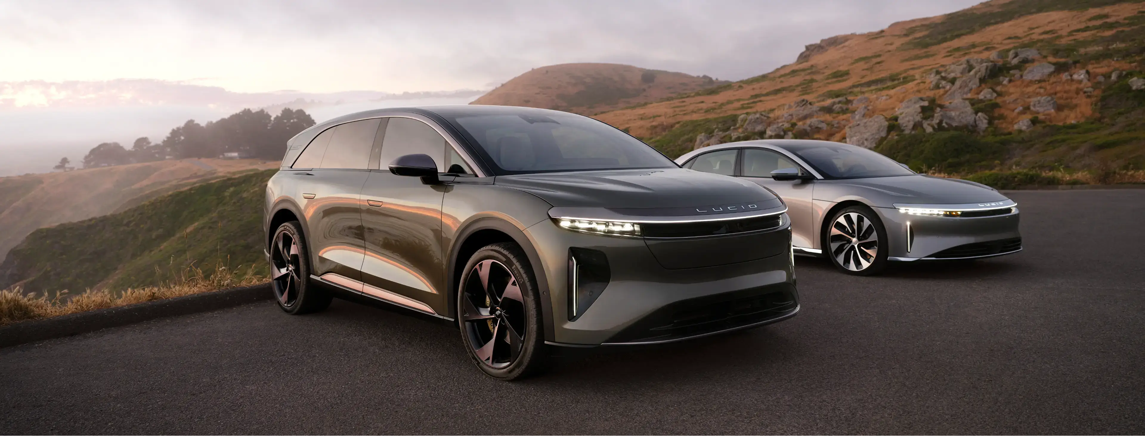Lucid Gravity and Lucid Air parked on a mountain road at dusk, with rugged hills in the background.