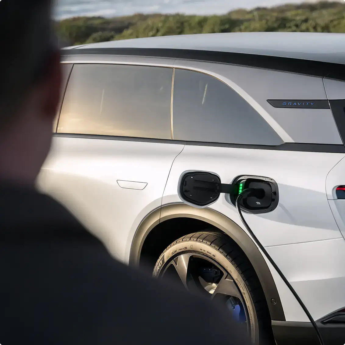 A person watches a white Lucid Gravity electric SUV being charged, with a green charging indicator light visible.
