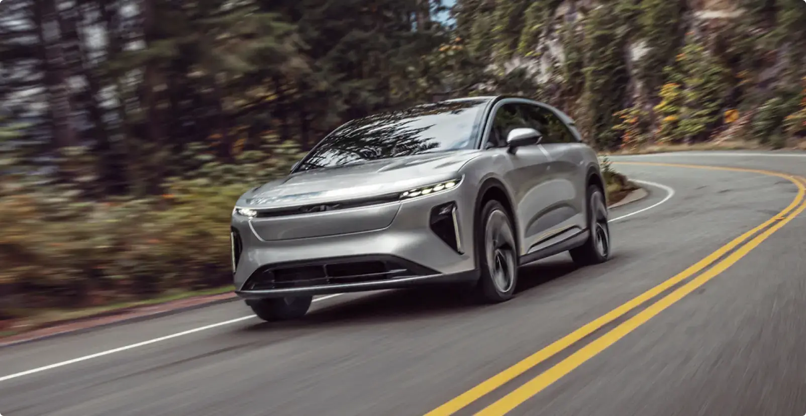 Lucid Gravity driving on a winding forest road with headlights on, motion blur suggesting speed.