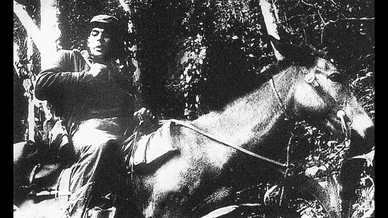 Schwarz-Weiß-Foto einer Person in Militärkleidung, die auf einem Pferd sitzt, umgeben von dichtem Laub und Bäumen. Das Bild hat eine körnige Textur, was darauf hindeutet, dass es historisch ist.