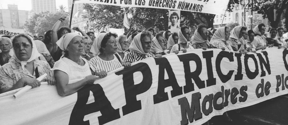 Eine Gruppe von Frauen mit weißen Kopftüchern marschiert mit einem großen Transparent mit der Aufschrift "Aparición Madres de Plaza" und fordert bei einem öffentlichen Protest Menschenrechte. Im Hintergrund sind Bäume und weitere Transparente zu sehen.