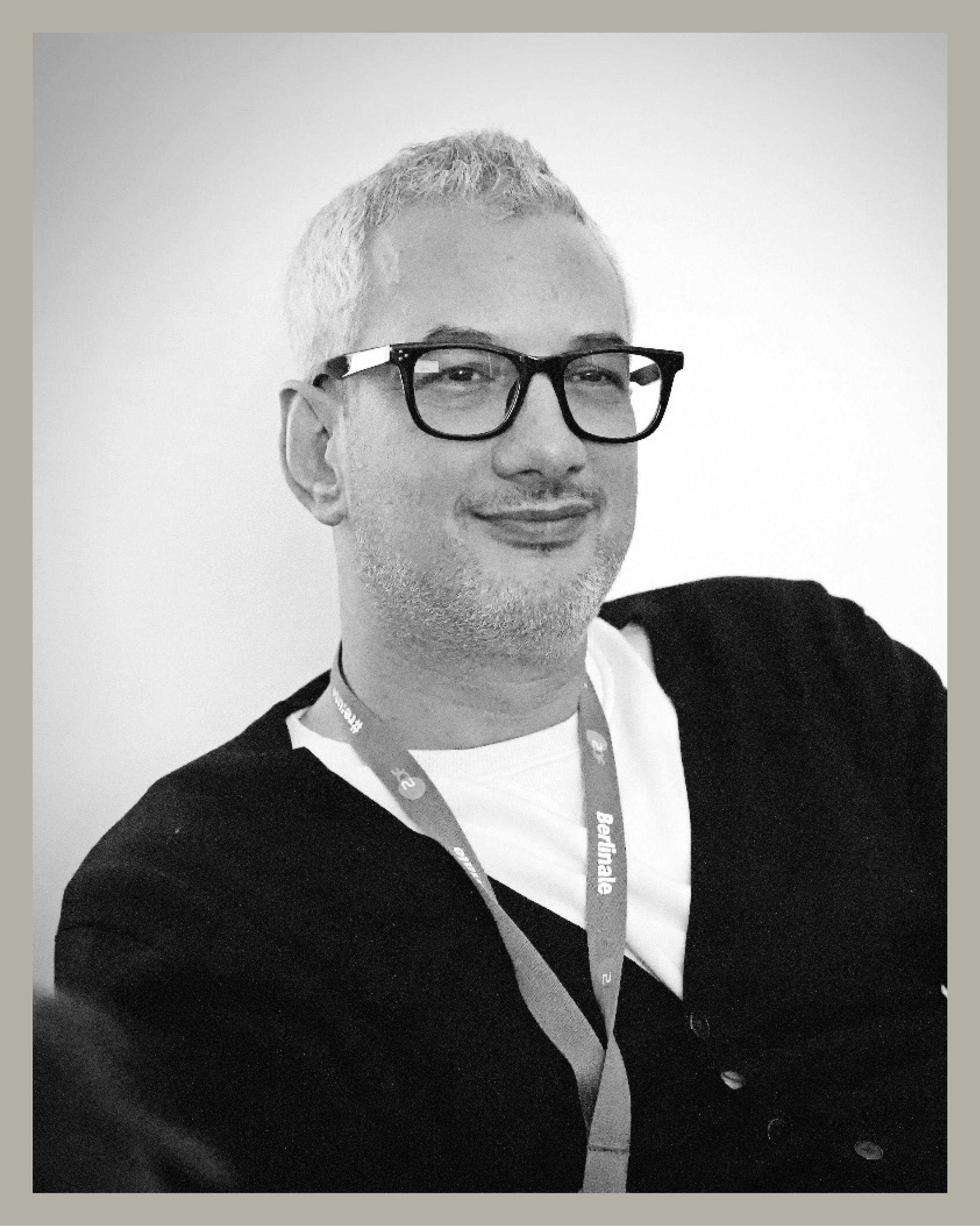 Ein Mann mit kurzen grauen Haaren, der eine schwarze Brille, eine dunkle Strickjacke über einem weißen Hemd und ein Schlüsselband um den Hals trägt, lächelt im Sitzen vor einem schlichten hellen Hintergrund. Das Bild ist in schwarz-weiß gehalten.