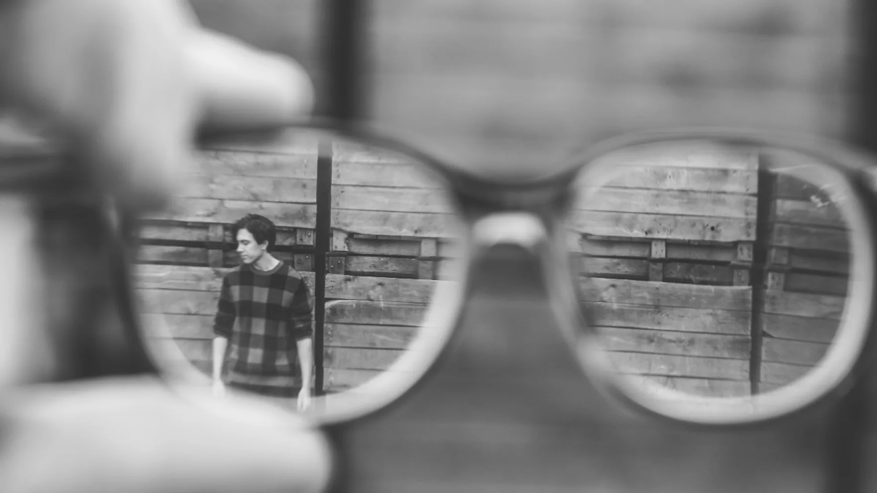 Photo by Joanna Nix on Unsplash. Image description: Black and white photo taken through a pair of glasses held up to the camera, of a man standing against a wall in the distance. The image is out of focus.