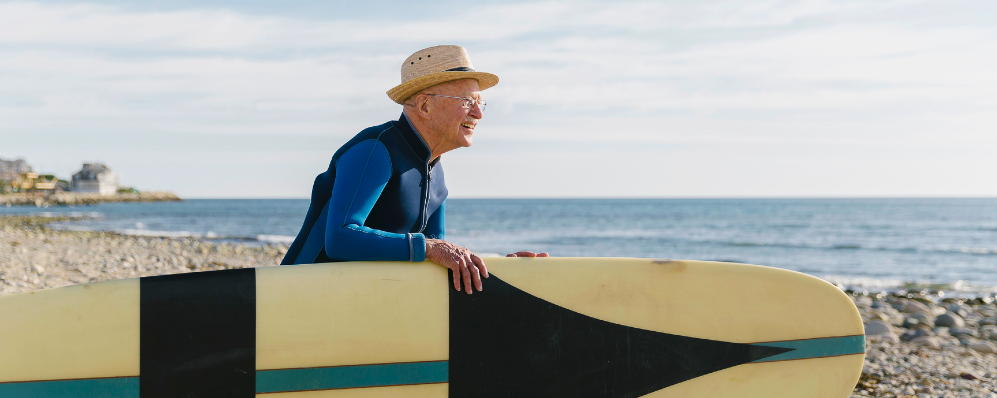 An elderly man walks to the beach with a surfboard.