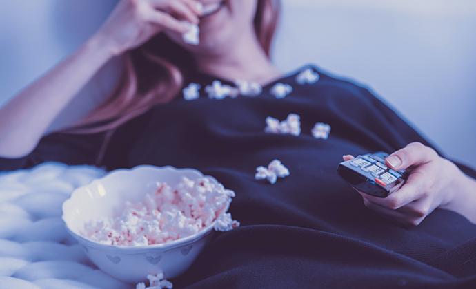 An image of a person laying down on their couch, eating popcorn, and watching television.