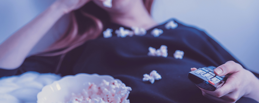 An image of a person laying down on their couch, eating popcorn, and watching television.