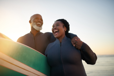 A couple laughs together holding a surfboard between them with the ocean in the background. 