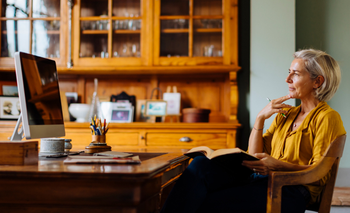 A middle-aged woman sits at a desk thinking with an open book in her lap.