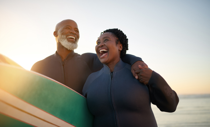 A couple laughs together holding a surfboard between them with the ocean in the background. 