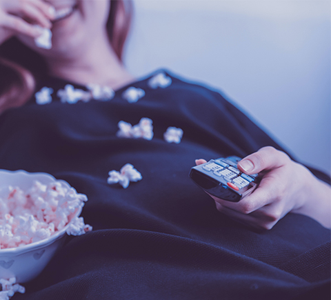 An image of a person laying down on their couch, eating popcorn, and watching television.
