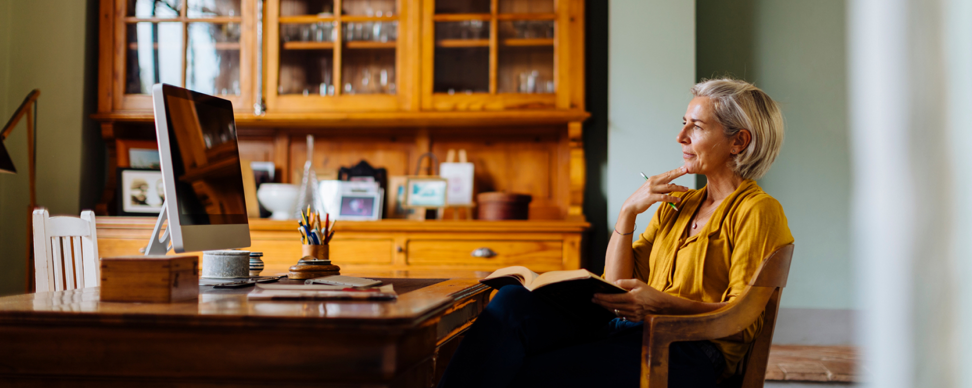 A middle-aged woman sits at a desk thinking with an open book in her lap.