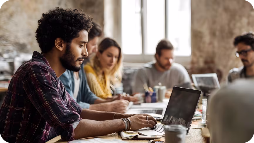Man sitting at a table working on his laptop with people around. 