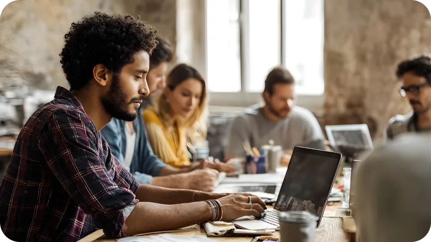 Man sitting at a table working on his laptop with people around.