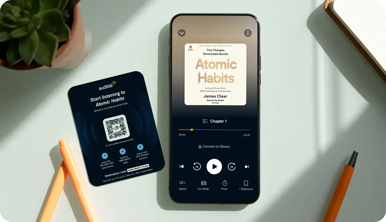 A smartphone siting on a desk with an Audible QR code card next to it.