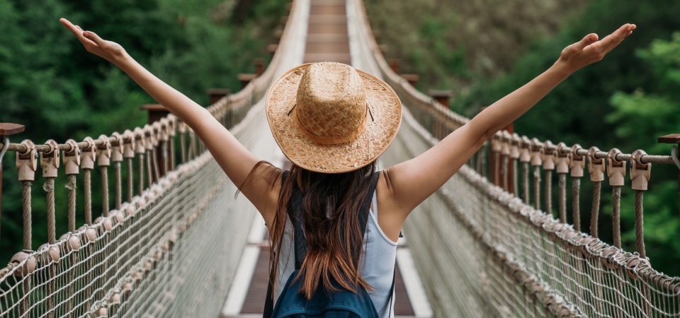 Ragazza su ponte tibetano. E' di spalle con le braccia alzate, indossa un cappello di paglia e uno zainetto. Il ponte si trova su una foresta tropicale