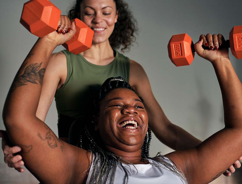 Bild: Eine Frau hält im Lady Gym in jeder Hand eine Kurzhantel. Hinter ihr steht eine weitere Frau und unterstützt sie beim Training.