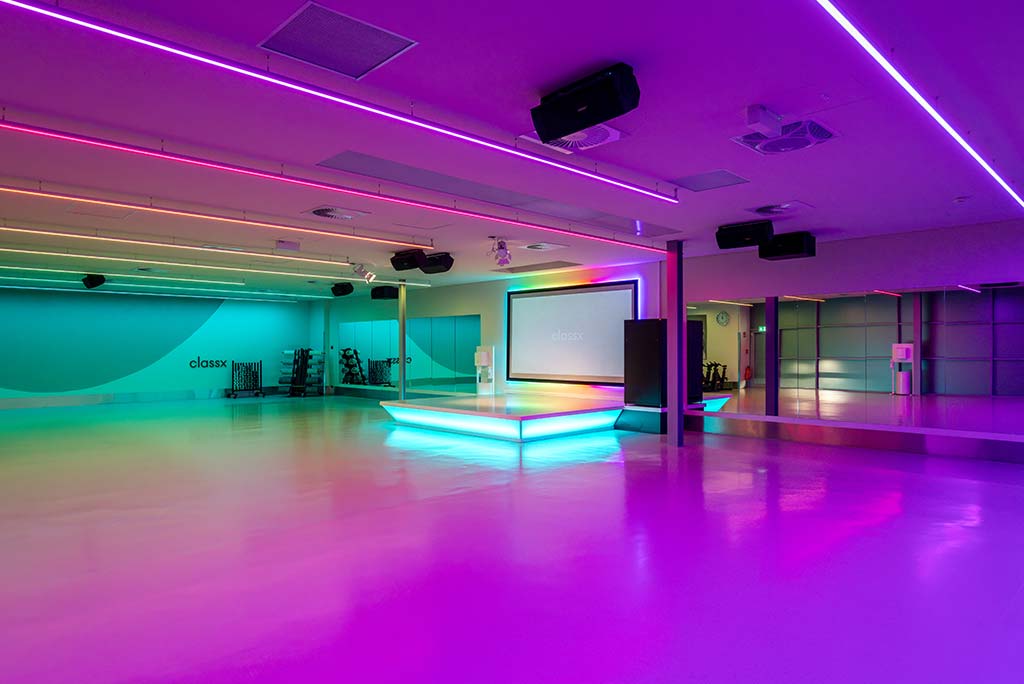Großer, offener Kursraum im FitX Fitnessstudio Neuss-Innenstadt. An der hinteren Wand befindet sich eine erhöhte Bühne, dahinter eine Leinwand. Links und rechts davon sind Spiegelwände.