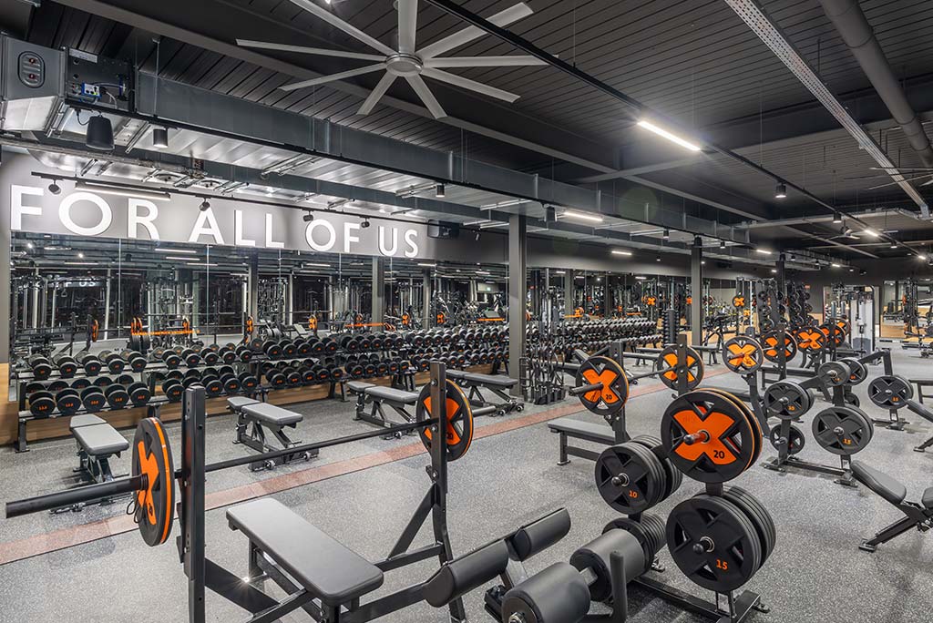 Weitläufiger Freihantelbereich mit verschiedenen Kraftgeräten im FitX Fitnessstudio Wuppertal-Elberfeld. An der Seite sind Kurzhanteln vor einer Spiegelwand aufgereiht. An der oberen Wand prangt der Schriftzug „FOR ALL OF US“.