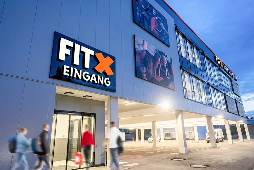 Bild: Außenansicht eines FitX Fitnessstudios. Ein großes FitX-Logo mit orangefarbenem X ist zu sehen. Das Gebäude befindet sich in einem urbanen Umfeld.