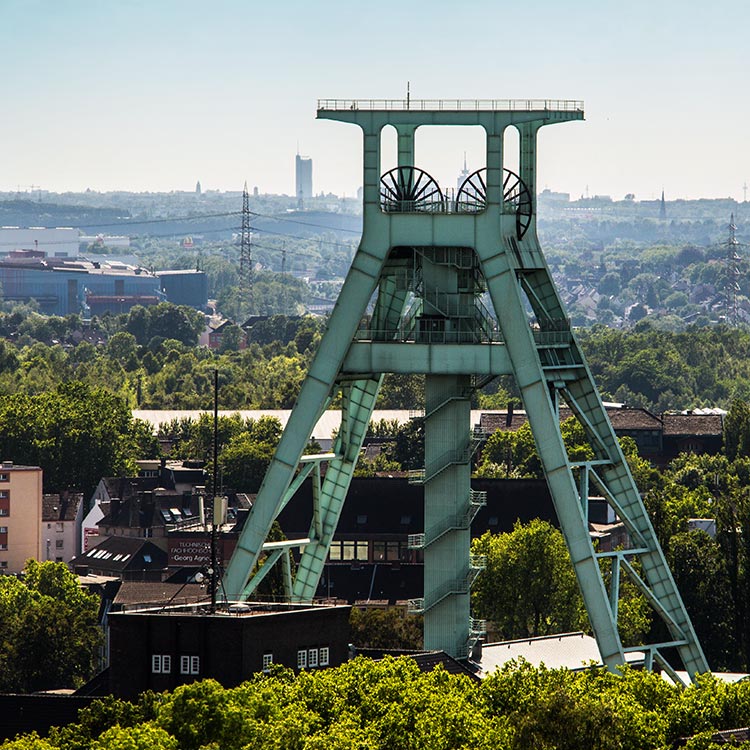 Grüner Förderturm des Deutschen Bergbau-Museums Bochum, umgeben von Bäumen und Stadtlandschaft im Hintergrund.